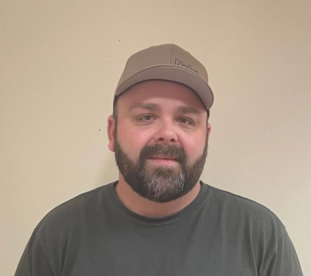 Bearded man wearing a tan baseball cap and dark shirt standing against a plain light wall.