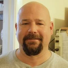 Bald man with goatee and light mustache, wearing a light gray shirt, smiling softly indoors.