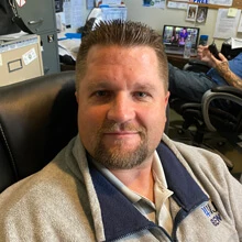 Man with short hair and beard sitting in office chair wearing a gray jacket.