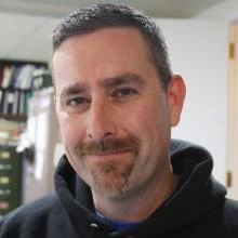 Man with short dark hair and beard smiling slightly indoors with bookshelves in background.
