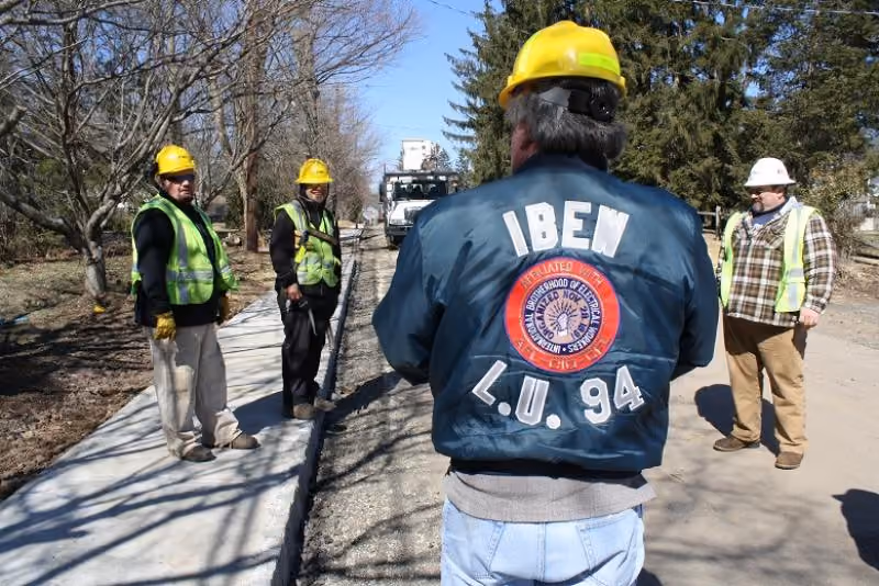 Four construction workers in safety gear standing beside a newly paved sidewalk on a clear day.