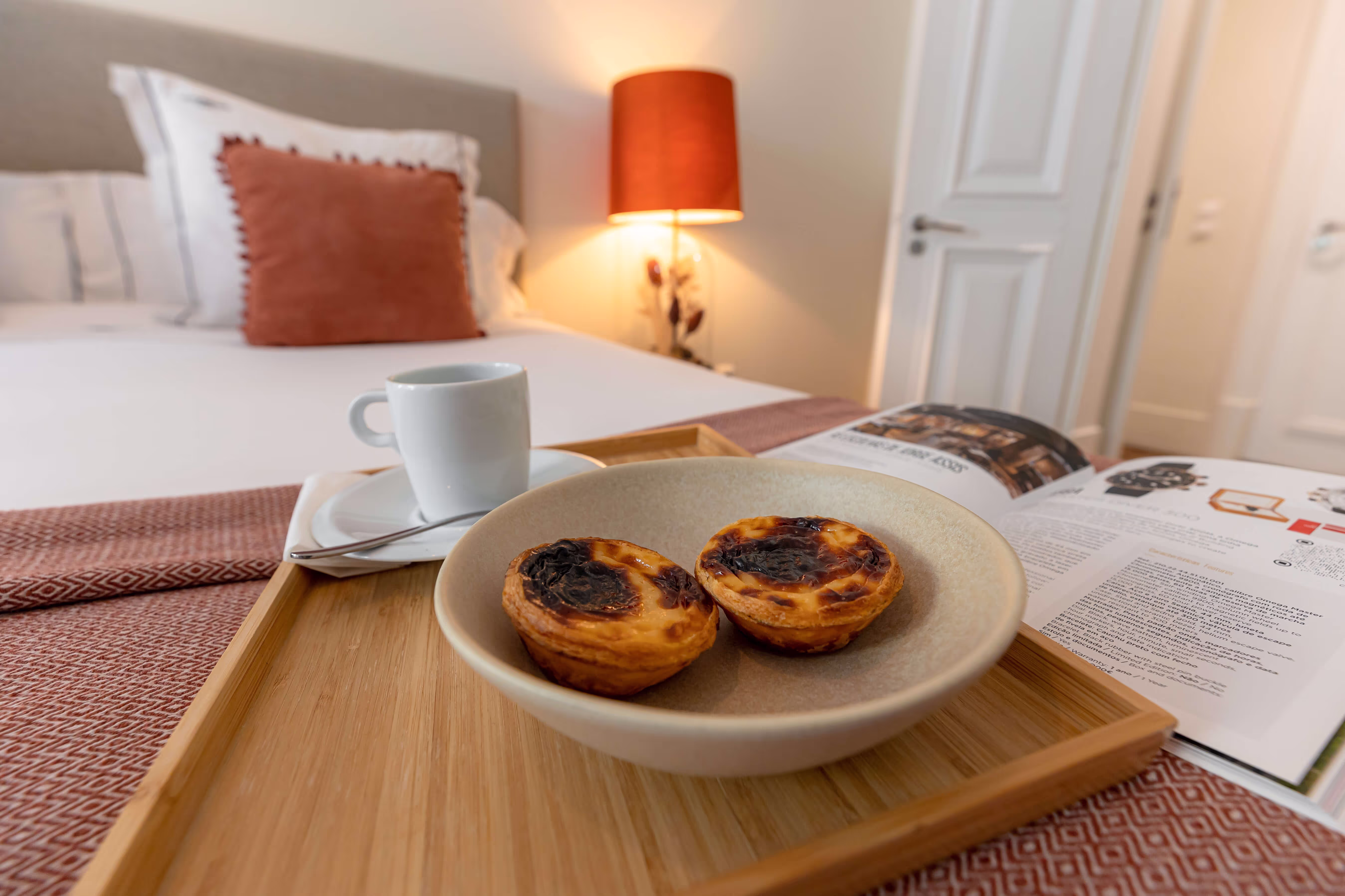Two Portuguese custard tarts on a plate with a coffee cup and magazine on a bed tray.