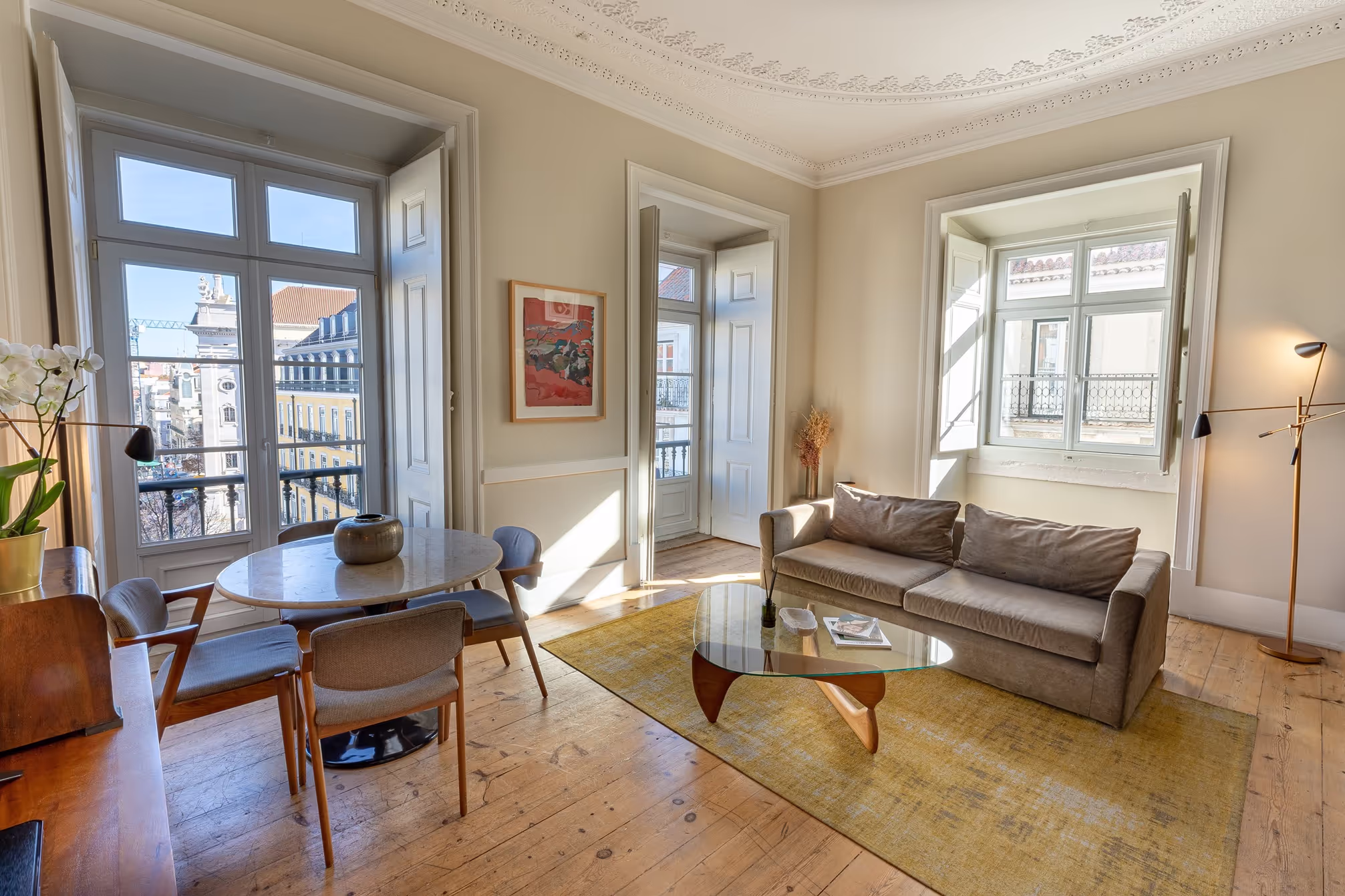 Bright living room with beige sofa, glass coffee table, round dining table with chairs, and large windows.