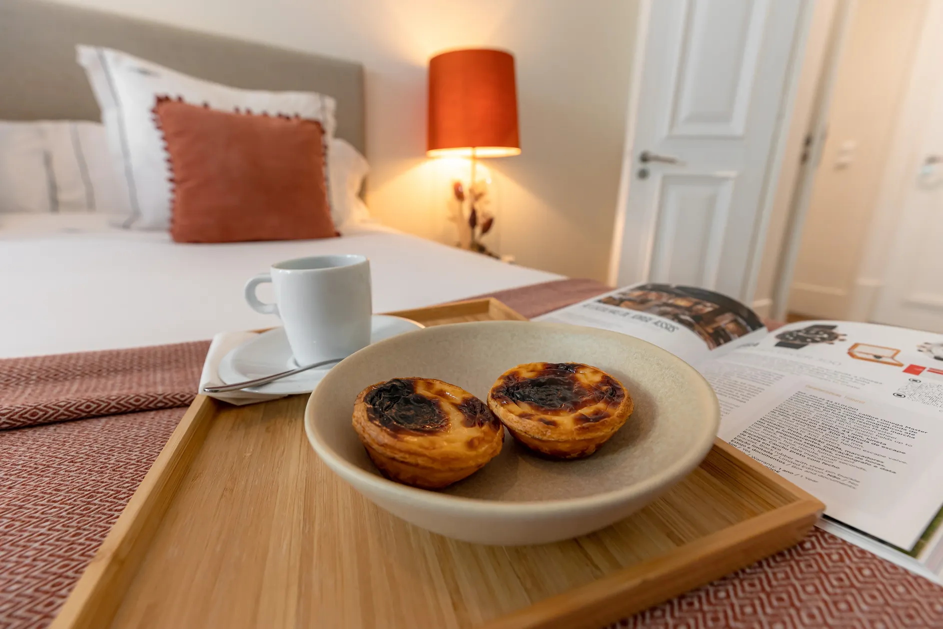 Two custard tarts on a plate with a cup of coffee and a magazine on a wooden tray in bed.
