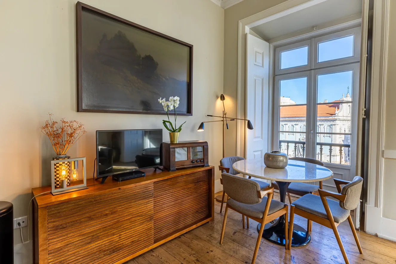 Cozy dining area with round table, four chairs, wooden sideboard, TV, vintage radio, and balcony view.