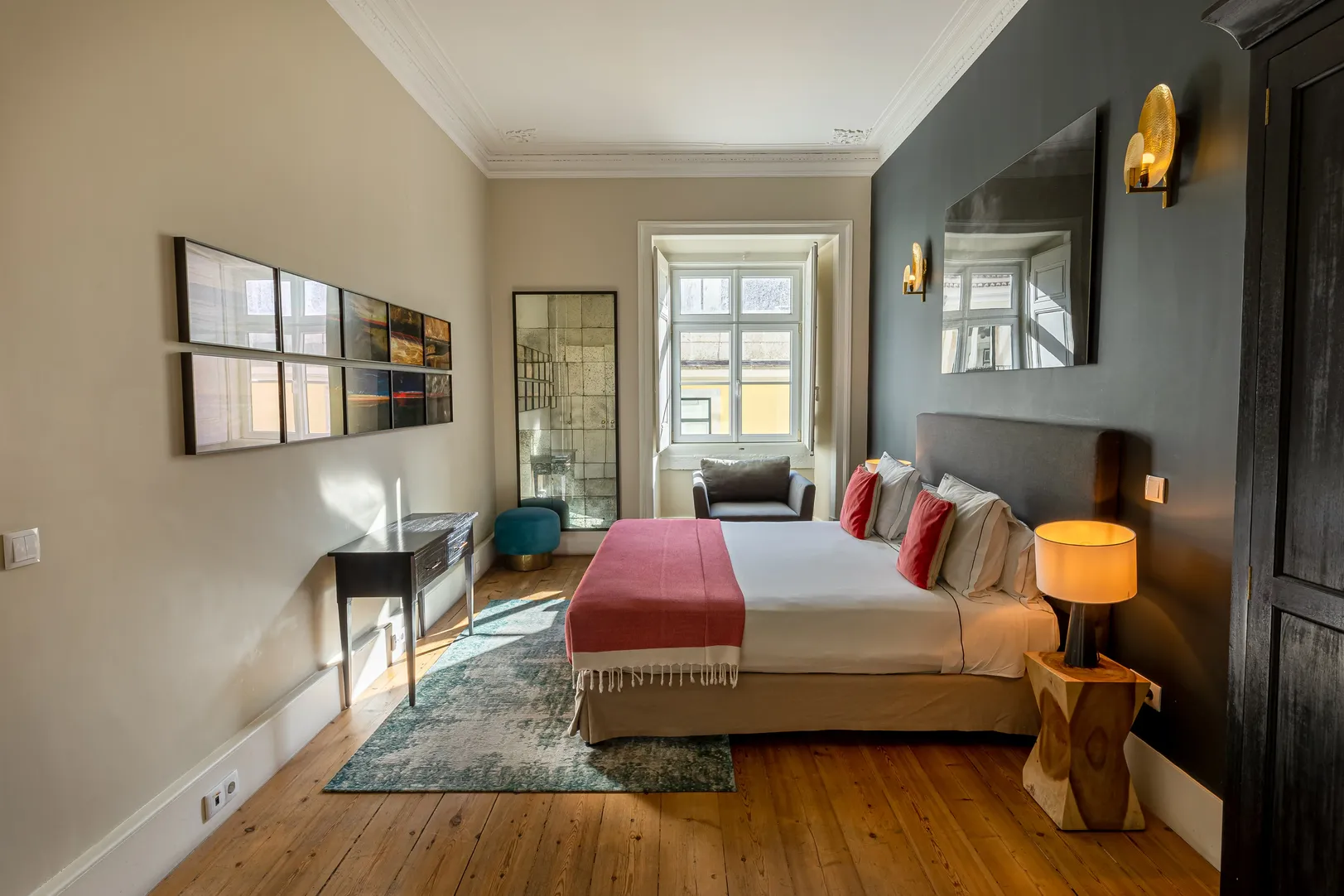 Modern bedroom with large bed, red pillows and throw, an armchair by the window, and wooden flooring.