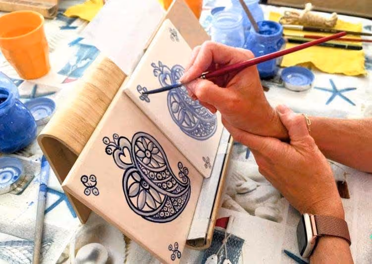 Person painting intricate blue floral patterns on tiles using a fine brush and holding a tile steady.