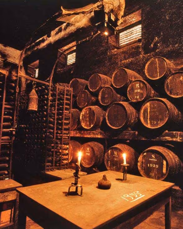 Dimly lit wine cellar with wooden barrels stacked, candles on a table, and wine racks in background.