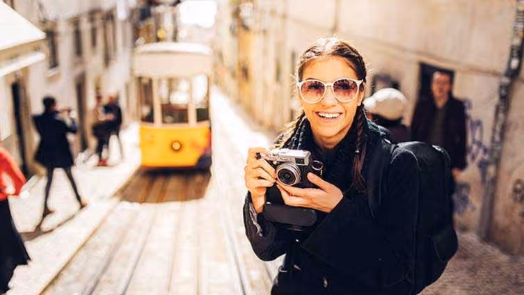 Smiling woman in sunglasses holding a camera on a street with a yellow tram in the background.