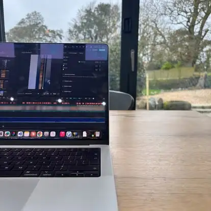 Part of a laptop with a video editing software open, on a wooden table by a window with trees outside.