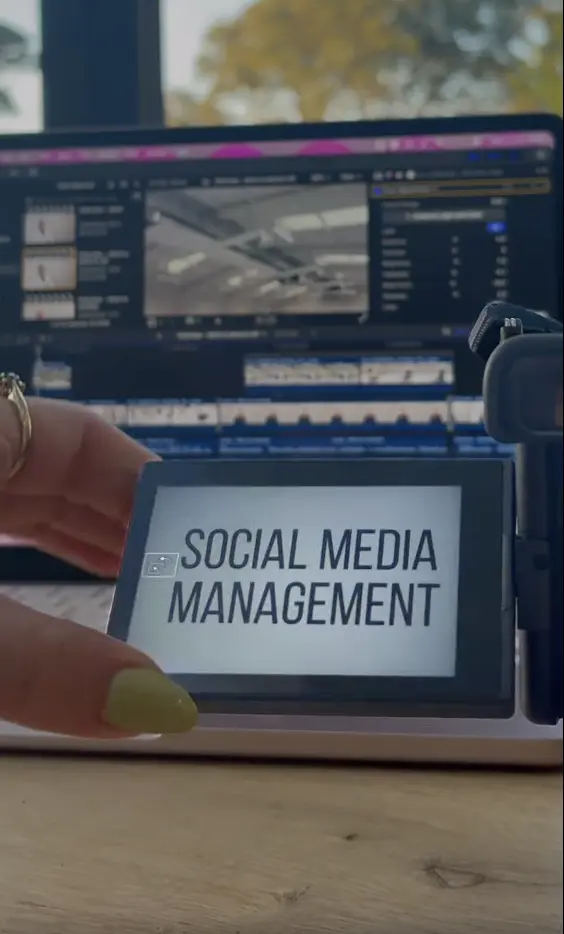 Hand holding a device screen displaying the text 'SOCIAL MEDIA MANAGEMENT' in front of a laptop.
