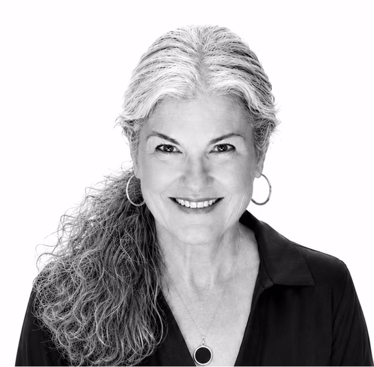 Smiling mature woman with gray hair in a ponytail, hoop earrings, and a dark top, on white background.
