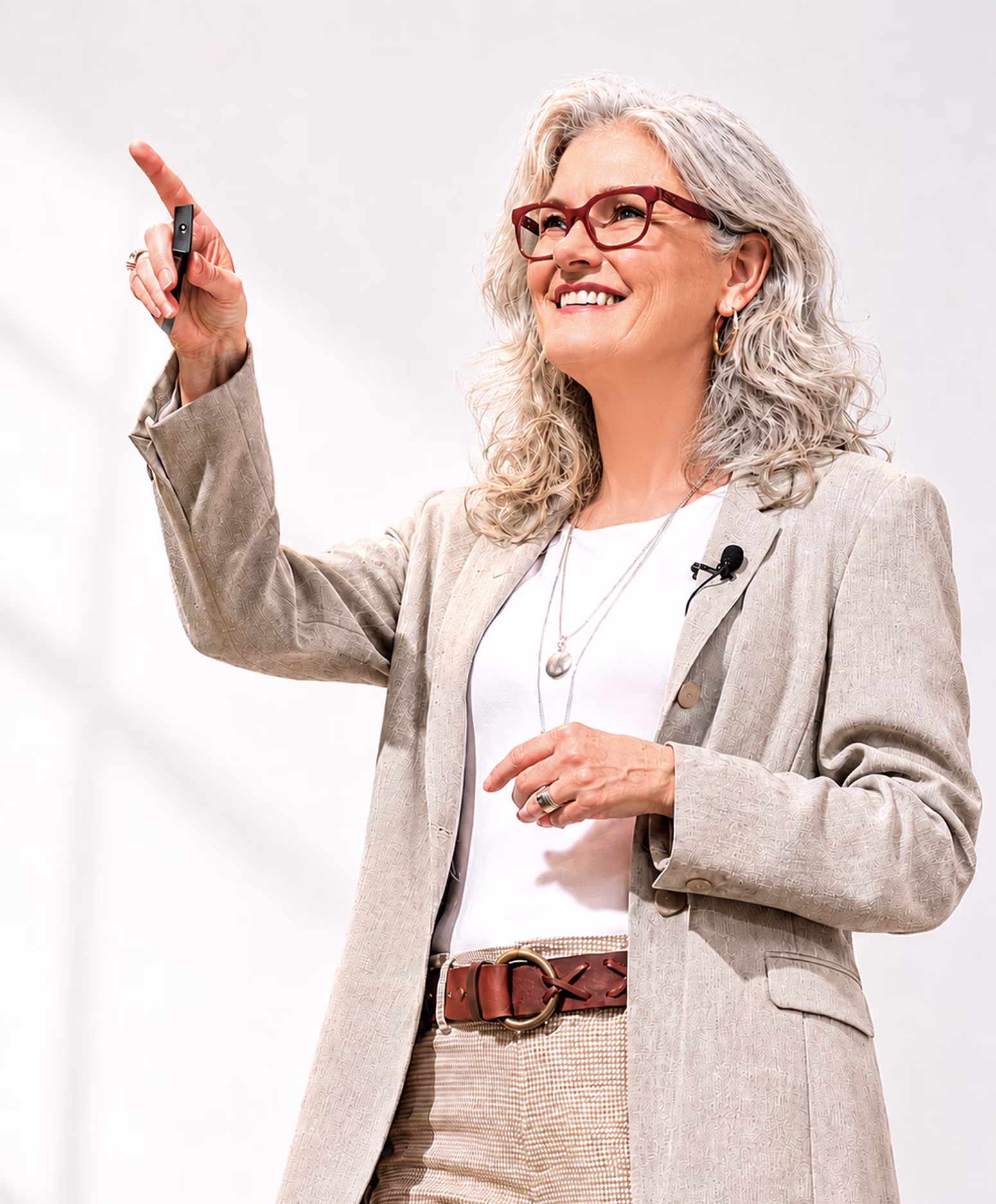 Smiling woman with gray curly hair and glasses pointing upward while wearing a beige blazer.