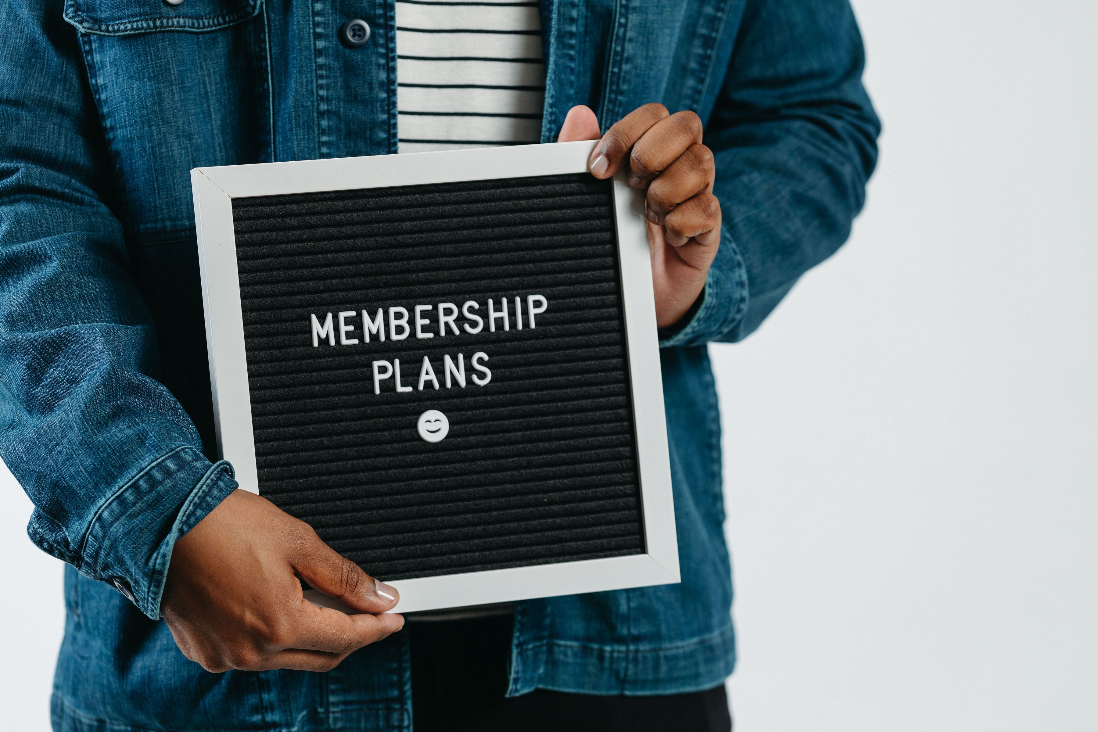 Person in denim jacket holding a black letterboard reading 'MEMBERSHIP PLANS' with a smiley emoji.