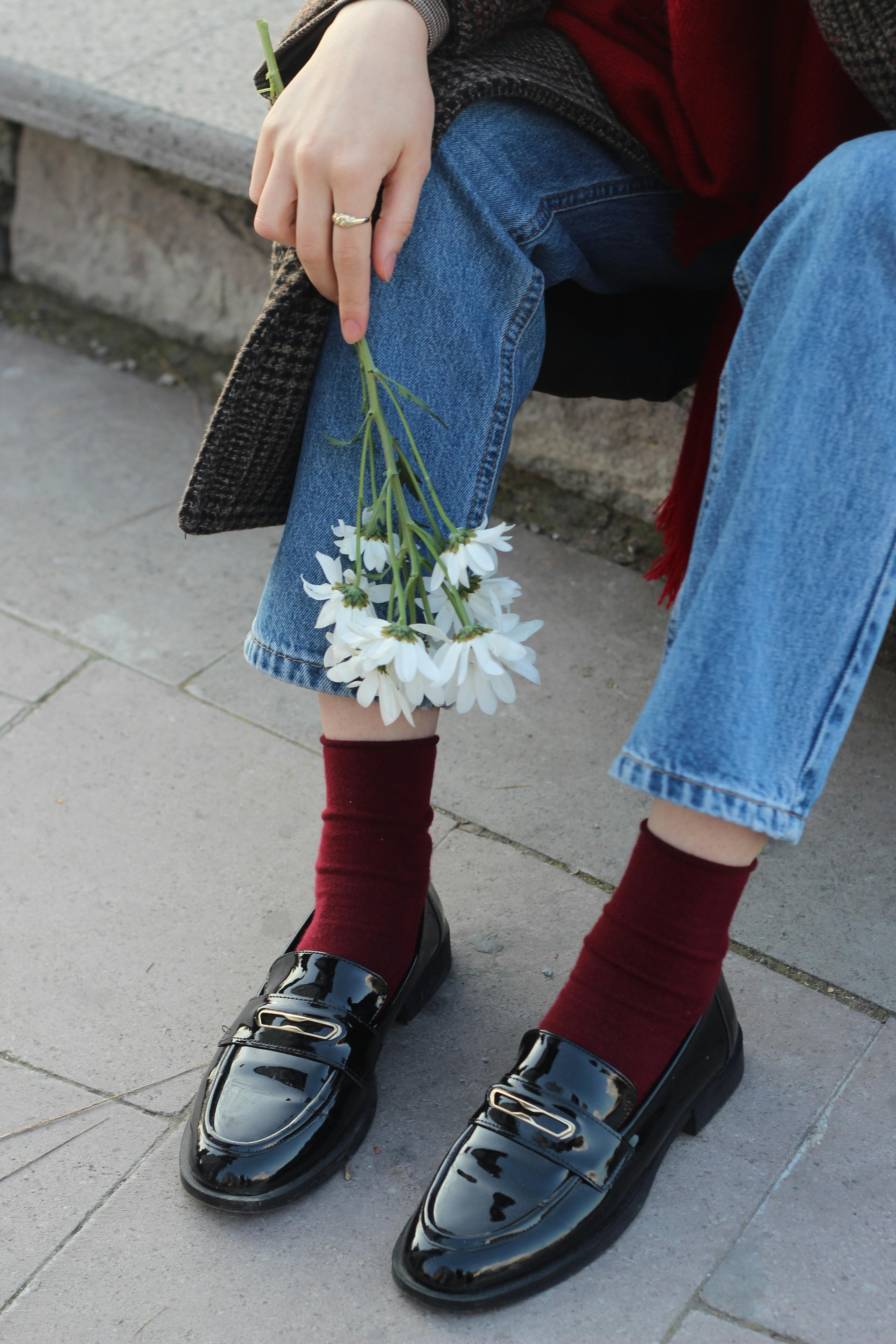 Person assise tenant un bouquet de marguerites blanches, portant des mocassins noirs et des chaussettes bordeaux.