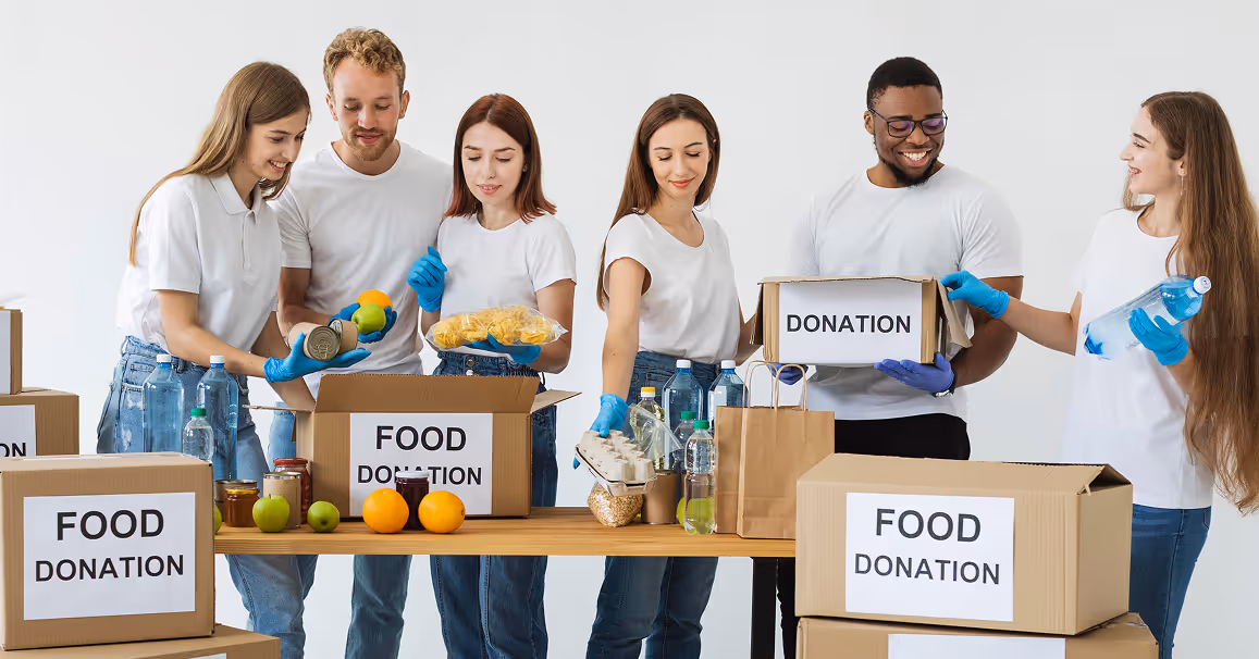 Group of young volunteers packing food and water bottles into boxes labeled 'Food Donation' and 'Donation' on a table.
