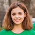Smiling woman with shoulder-length brown hair wearing a green top, standing outdoors with a blurred natural background.