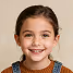 Smiling young girl with dark hair wearing denim overalls and a brown shirt against a beige background.
