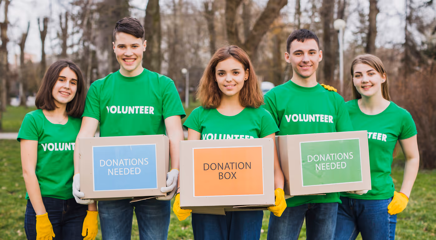 Five volunteers in green shirts holding donation boxes labeled 'Donations Needed' and 'Donation Box' outdoors.