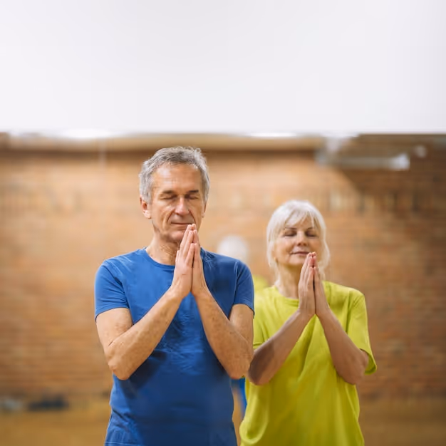 Older man and woman standing with eyes closed and hands pressed together in a prayer or meditation pose indoors.