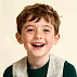 Smiling young boy with curly hair wearing a green vest and white shirt against a beige background.