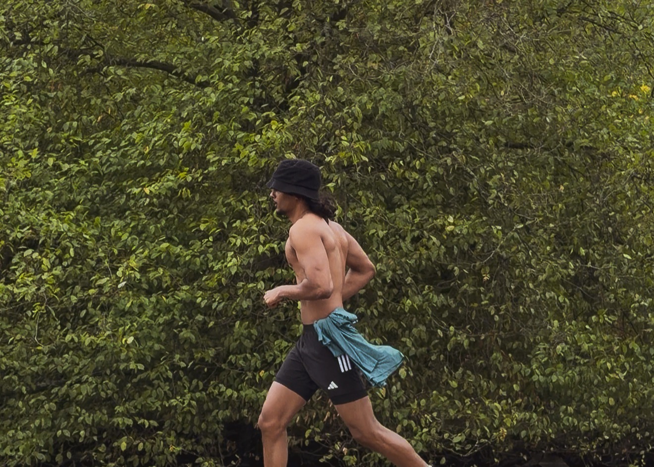Shirtless man running outdoors wearing black shorts and a black bucket hat, with a teal shirt tied around waist.