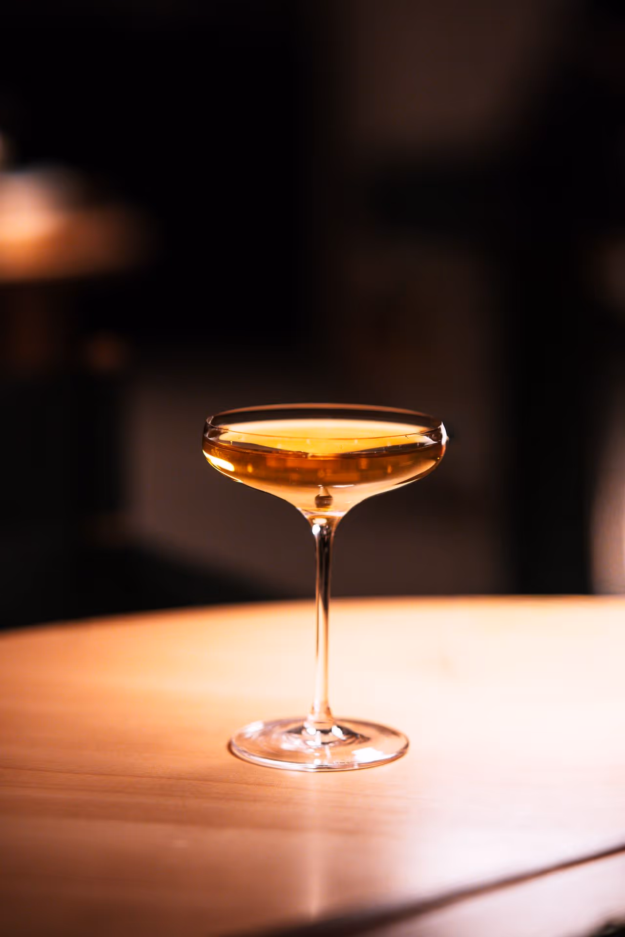 A coupe glass filled with a golden-colored cocktail on a wooden table in dim lighting.