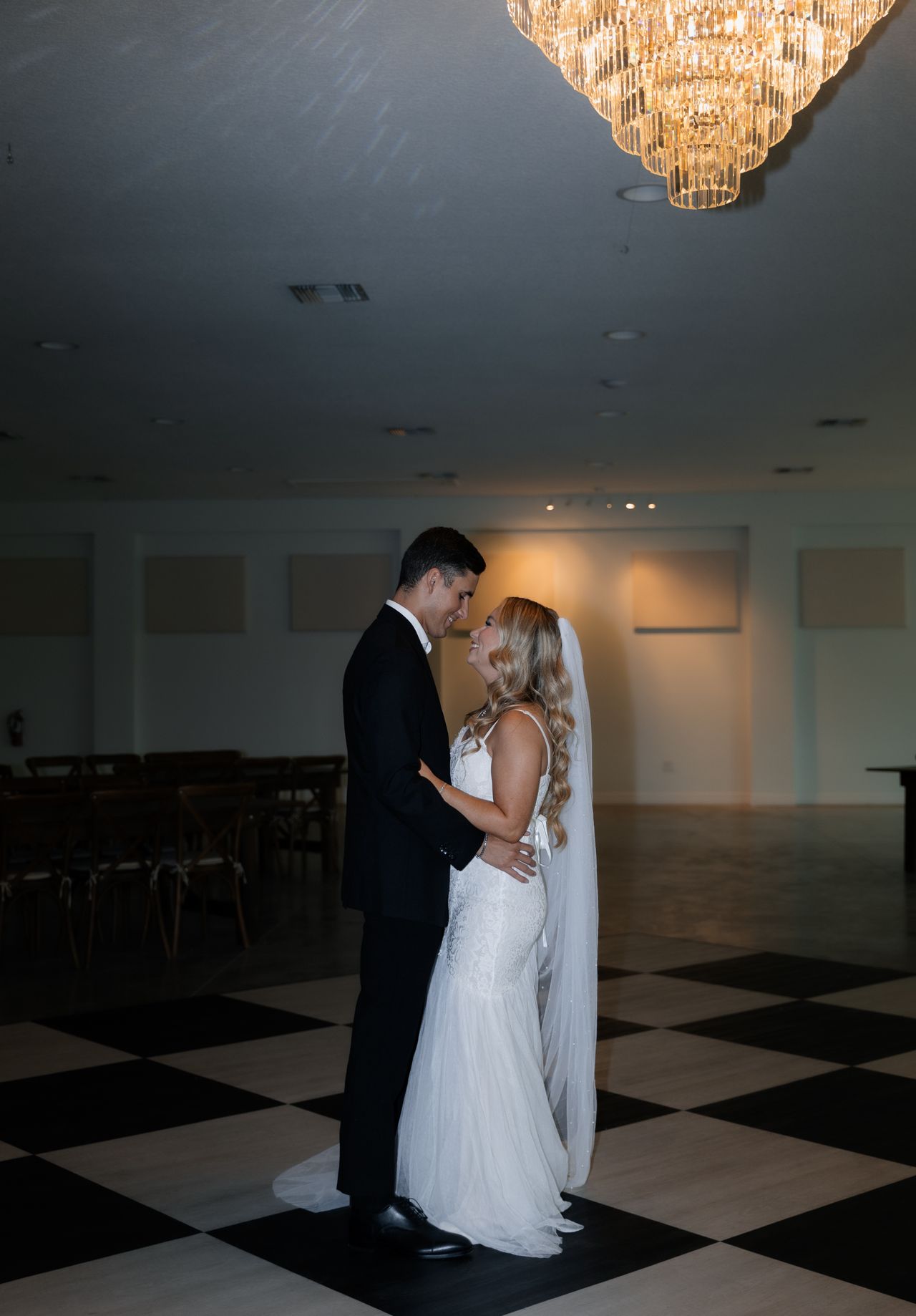 Bride and groom embracing on a checkered floor under a large chandelier in a dimly lit room.