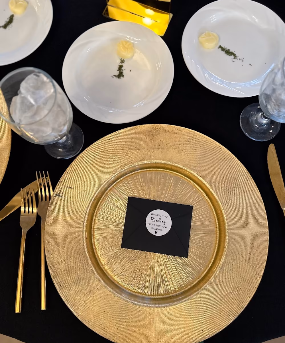 Elegant table setting with gold charger plate, gold utensils, black envelope, and plates with butter and herbs.