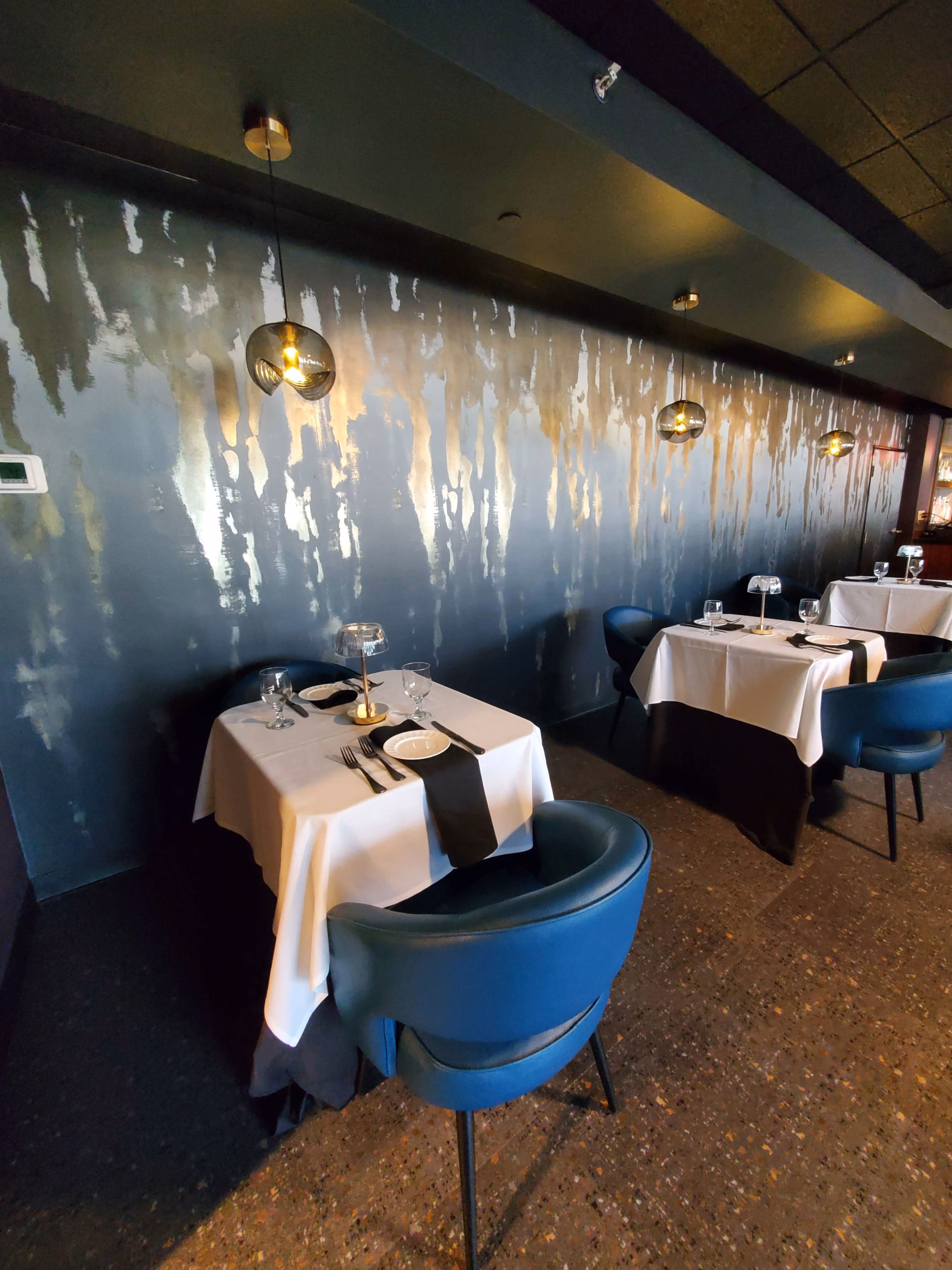 Elegant restaurant seating with white tablecloths, blue chairs, and modern pendant lights on textured wall at Isabella's Cocktail Lounge and Restaurant in Akron, OH.