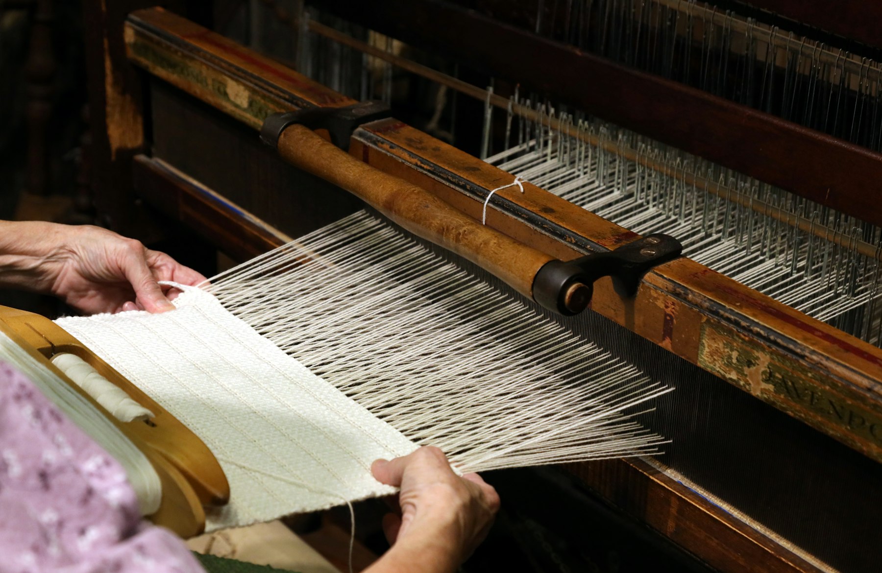 Hands guiding woven cloth through the warp threads of a wooden handloom
