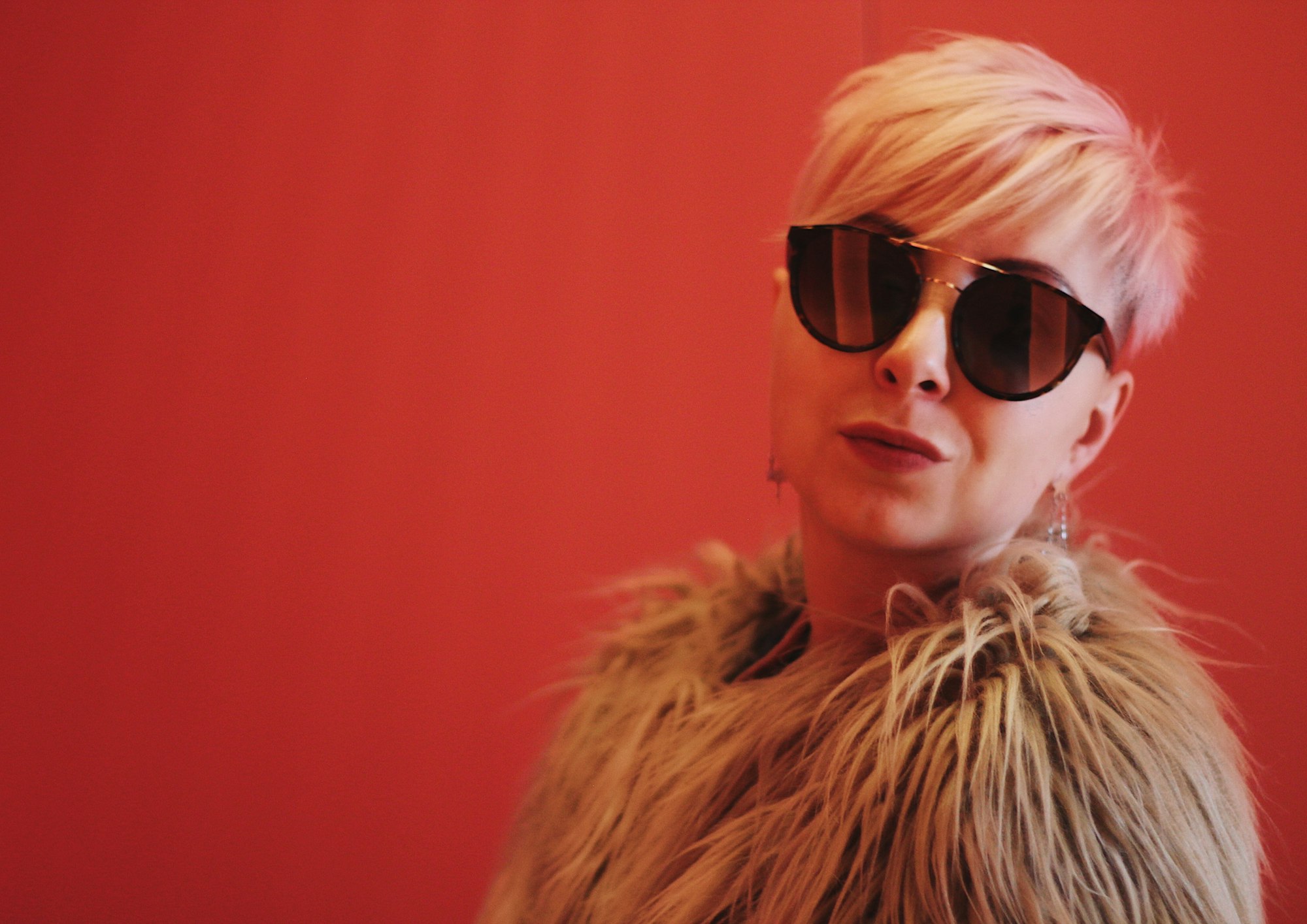 A woman with a pink-blonde pixie crop in round sunglasses and a shaggy faux-fur coat, chin raised against a bright red wall