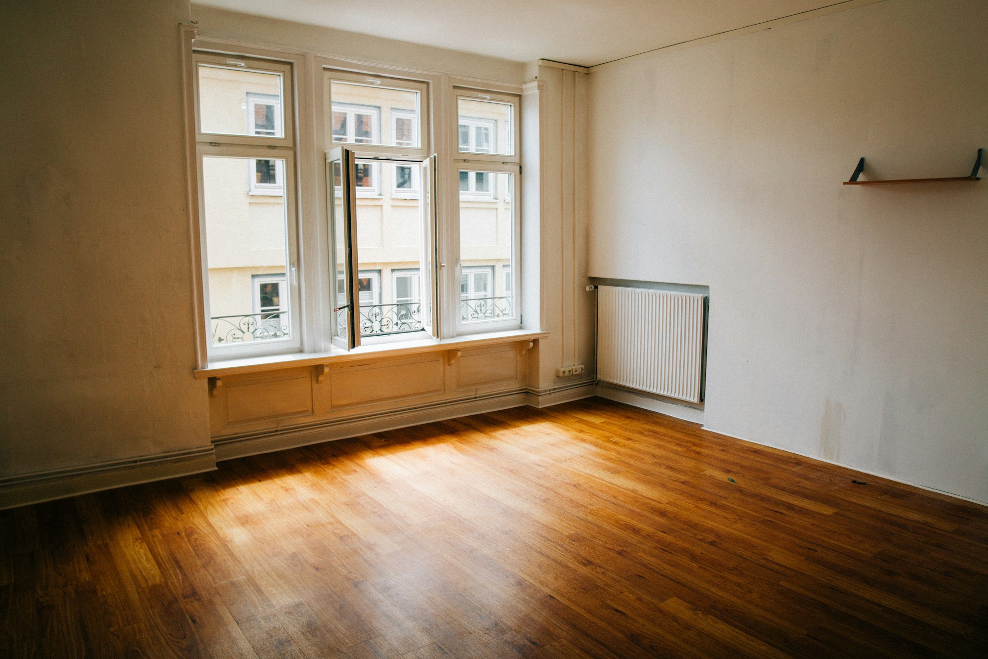 An emptied room with bare wooden floorboards, a tall sash window, a radiator and a single empty shelf on the wall