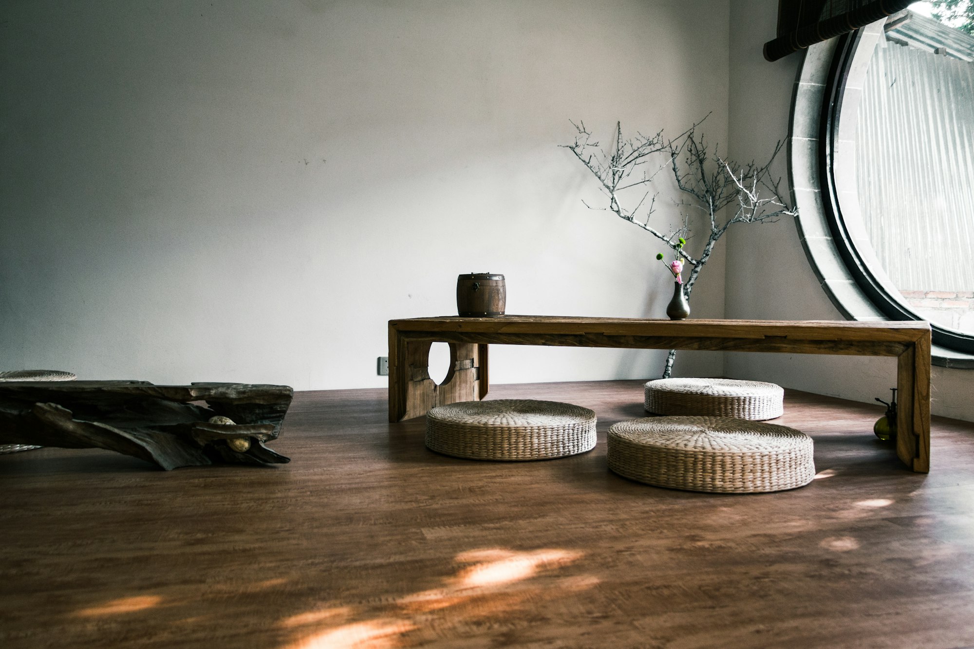 A minimal interior with a low wooden bench, woven floor cushions, a bare branch arrangement and a large round moon window