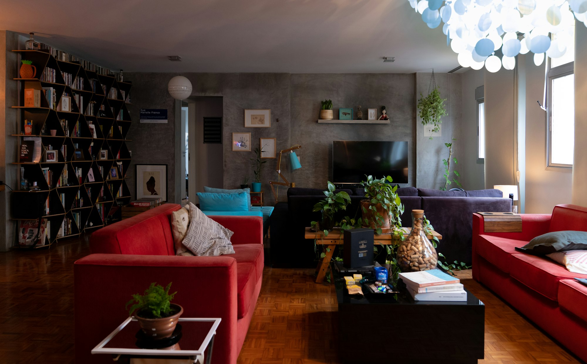 A living room with two deep red sofas facing a low black coffee table, beside a tall geometric bookshelf and a bare concrete wall