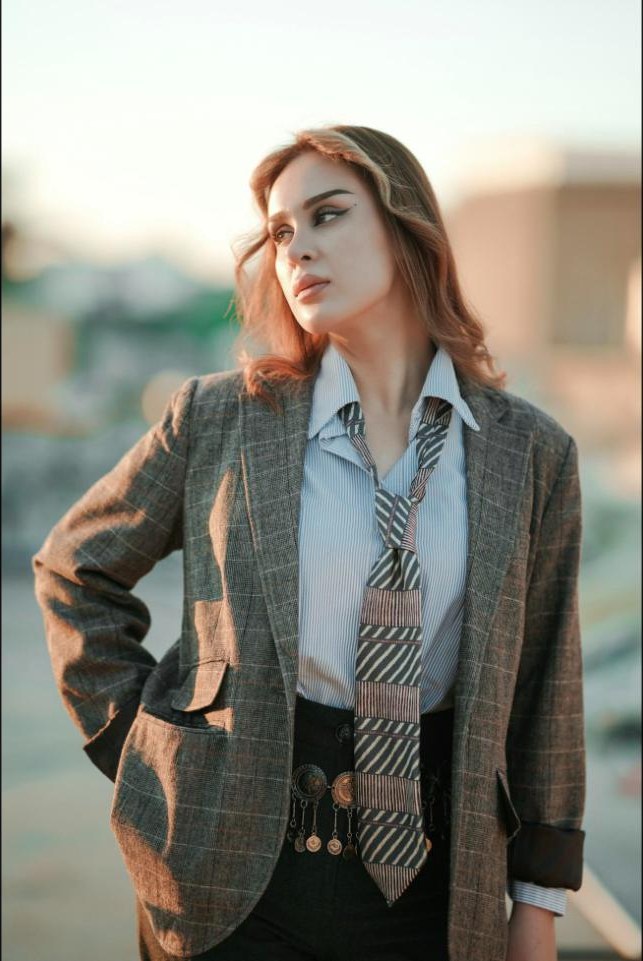 A woman in an oversized tweed blazer with a striped tie hanging loose over a shirt and belt