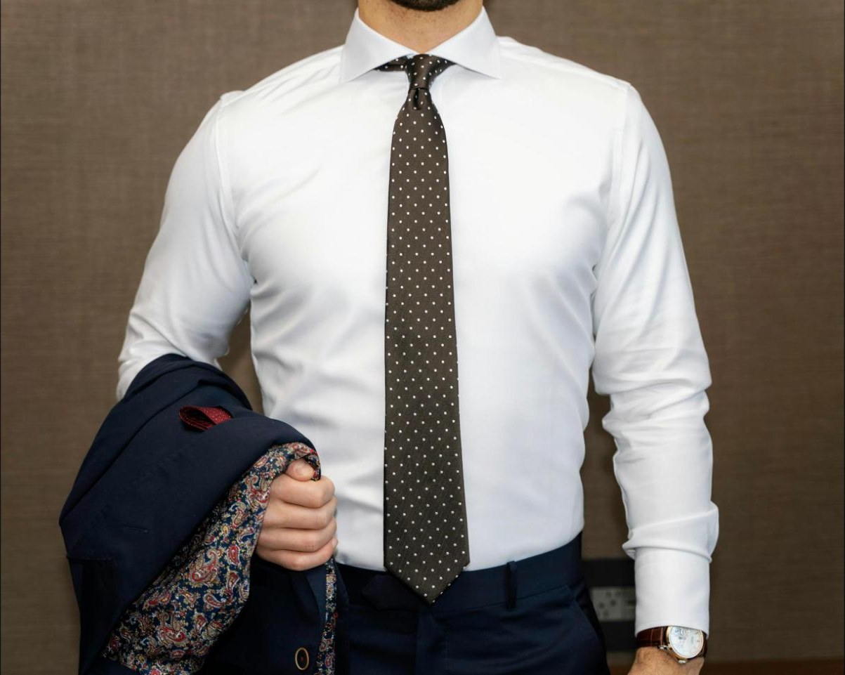 A dark spotted tie knotted over a white dress shirt, jacket carried over the arm