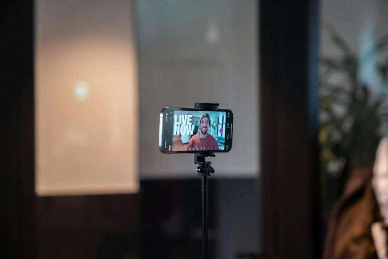 A smartphone on a tripod broadcasting a live stream, the screen reading LIVE NOW