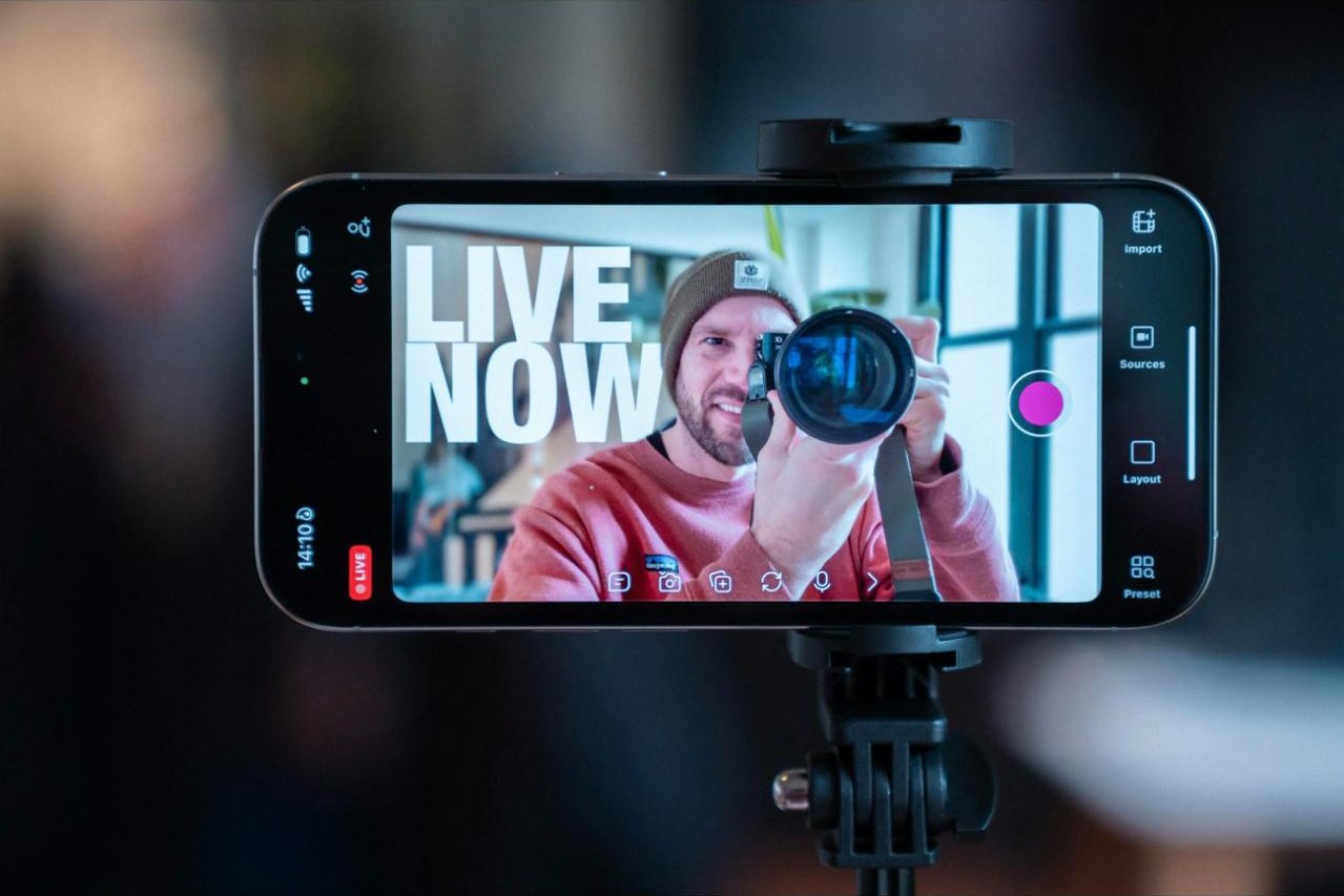 A phone on a tripod filming a person holding a camera, the screen reading LIVE NOW