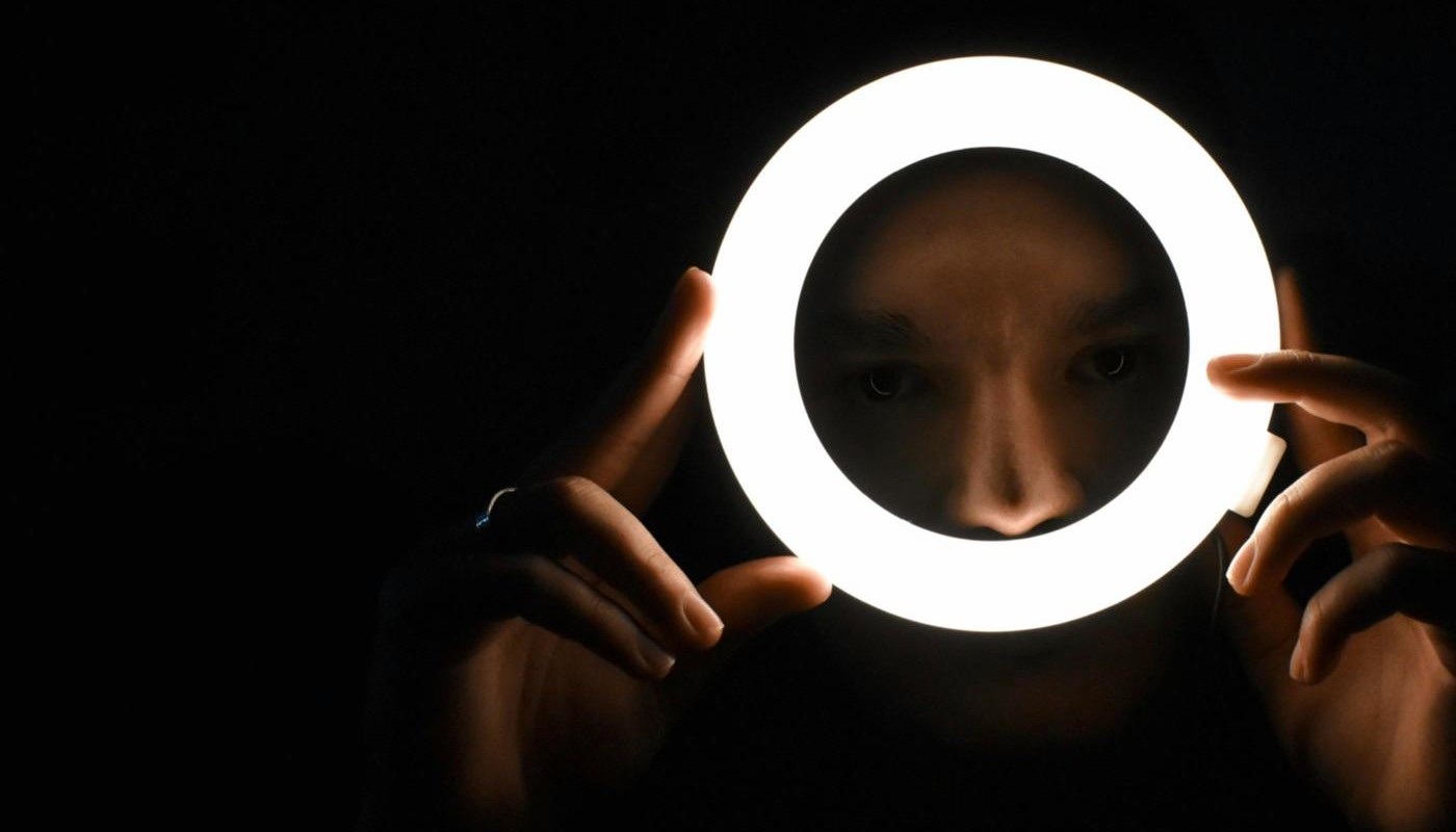 A face lit from the front, framed inside the circle of a ring light held up in the dark