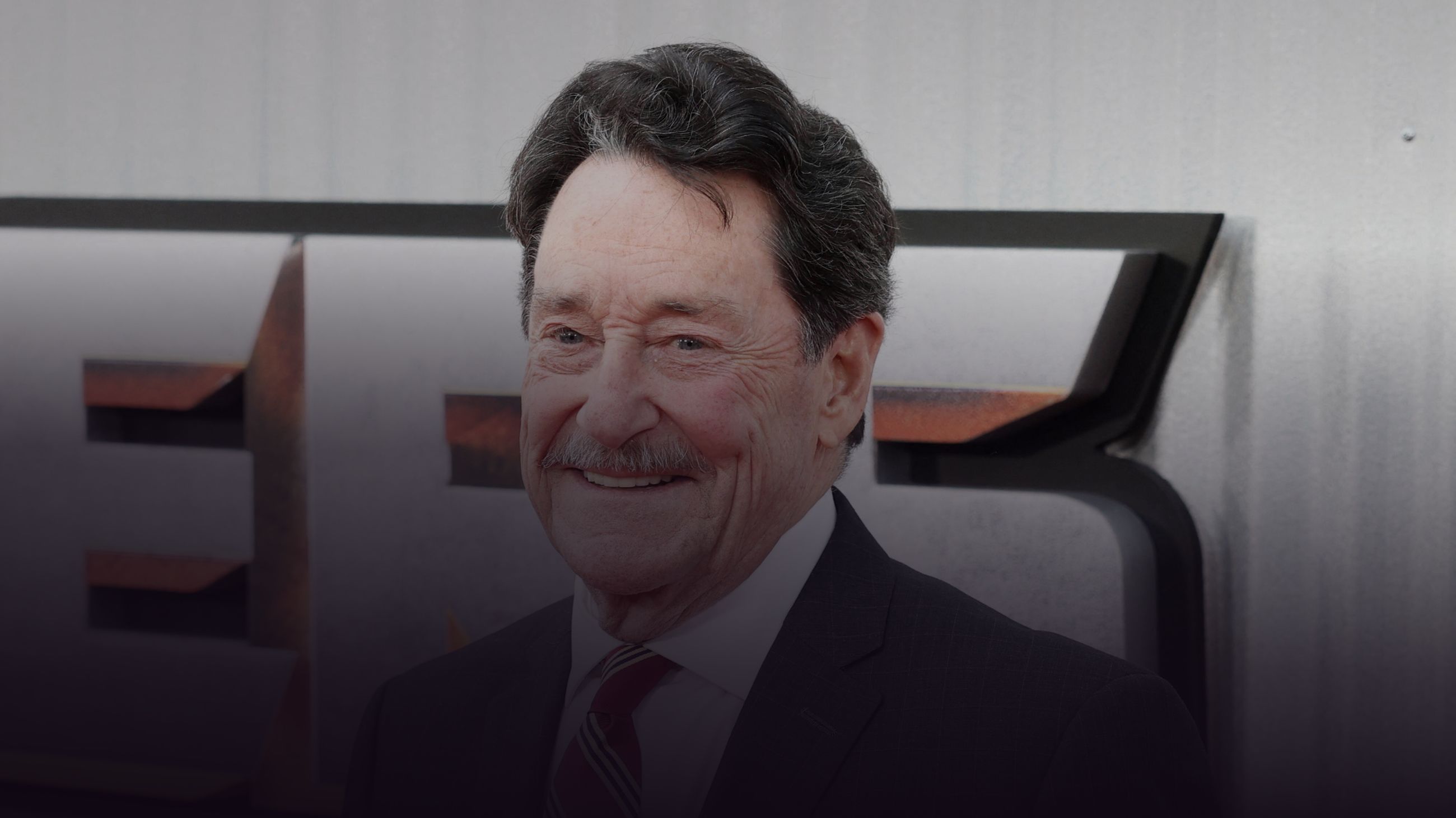 Peter Cullen, smiling, photographed in front of a Transformers logo wall at a film premiere.