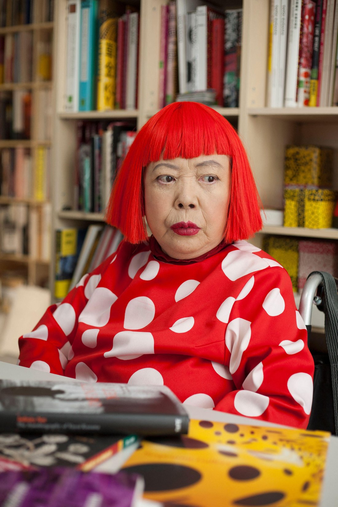 Yayoi Kusama in a red wig and red polka-dot dress, seated at a desk in her Tokyo studio