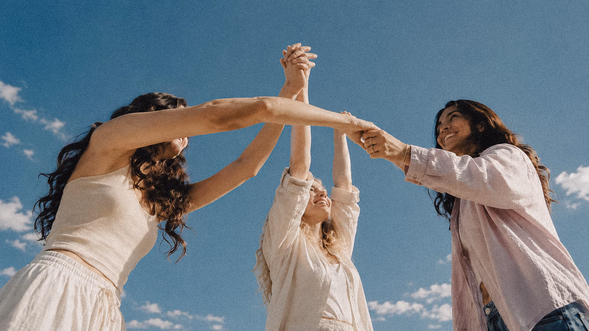 Three women laugh while holding raised hands against a blue sky
