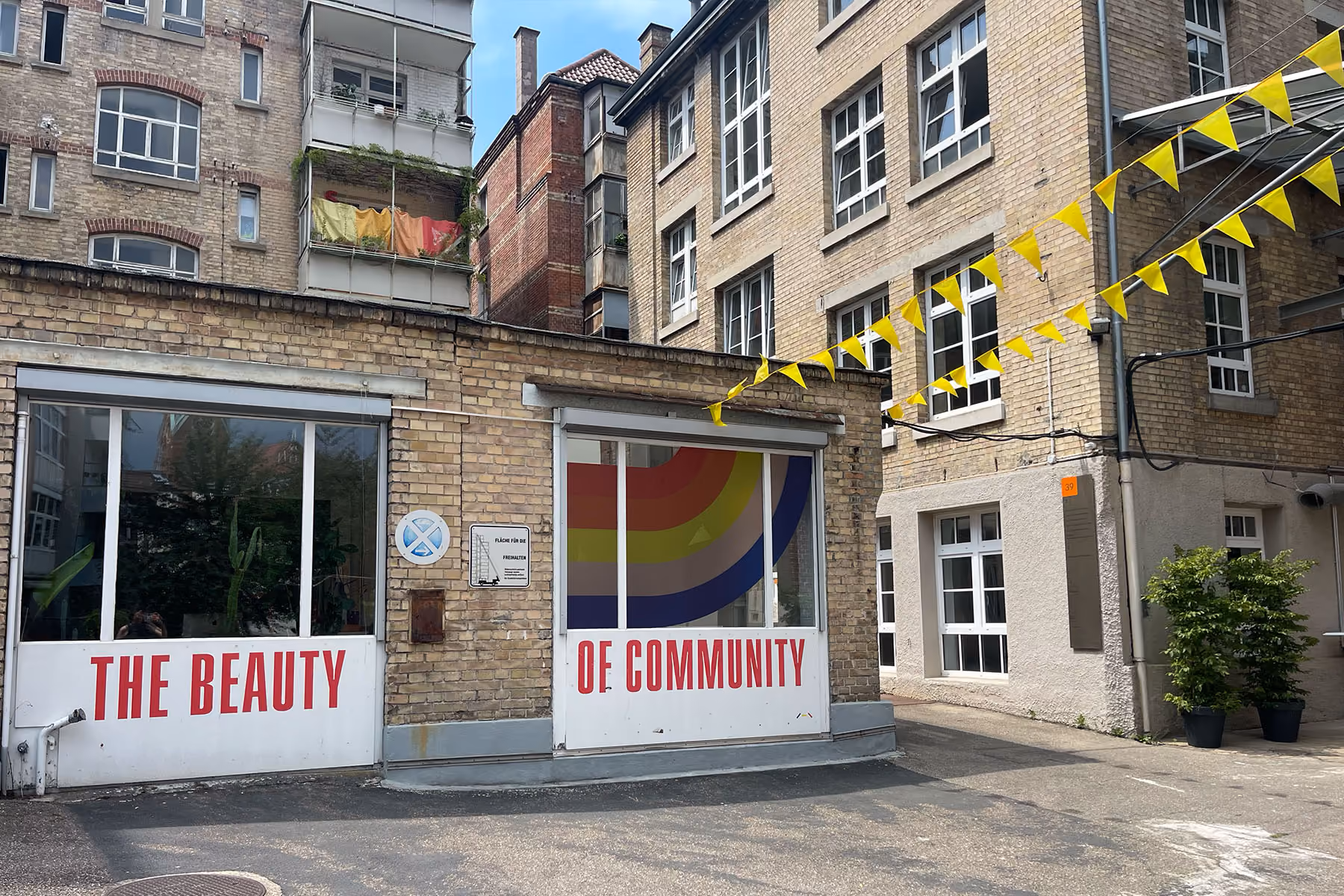 Gebäude mit Fenstern, zwei Türen mit Text ‚The Beauty‘ und ‚of Community‘ und gelben Wimpeln.