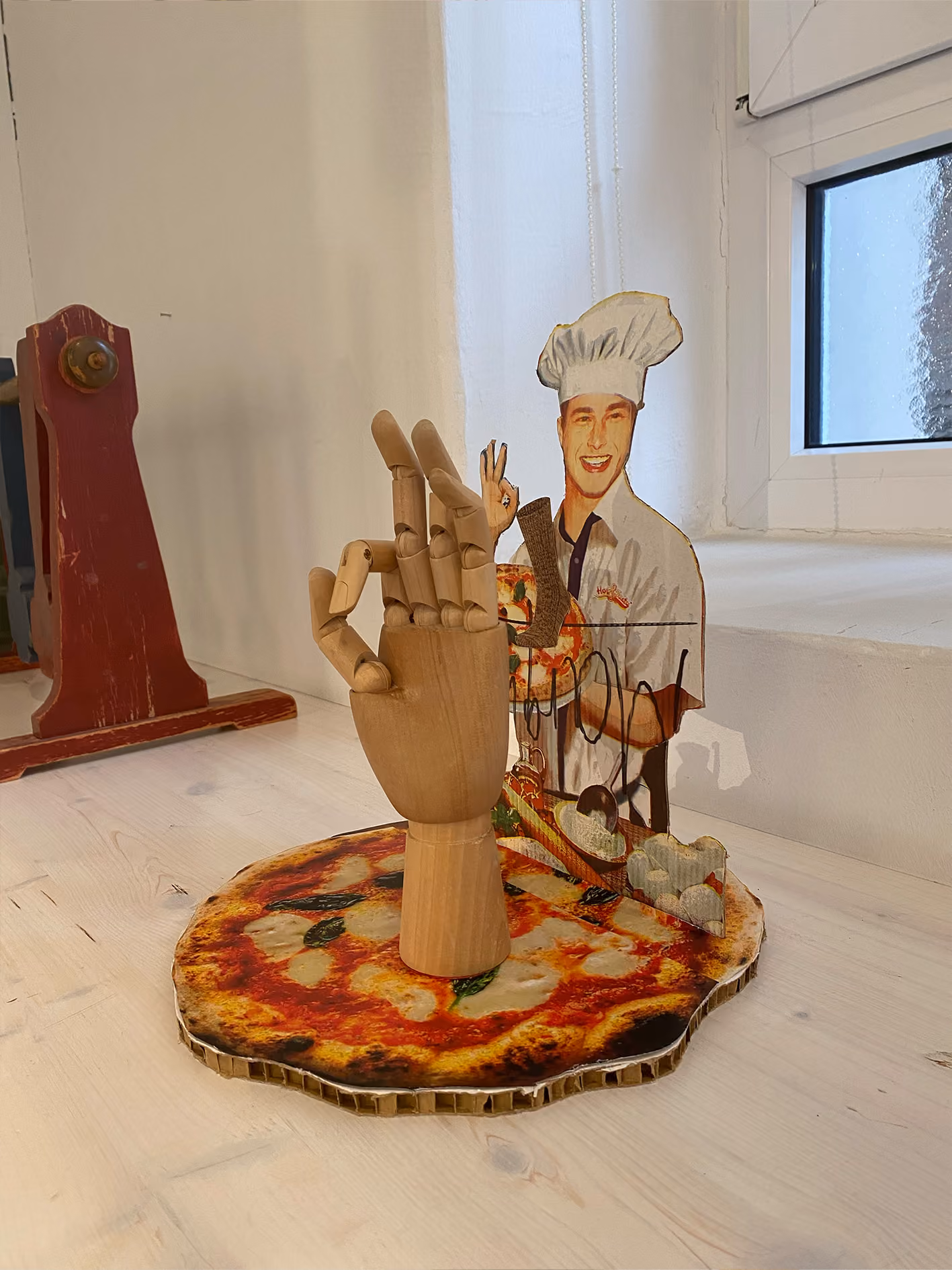 Hölzerne Gliederhand formt ein OK-Zeichen auf einer Fake-Pizza vor einer Pizzabäcker-Figur