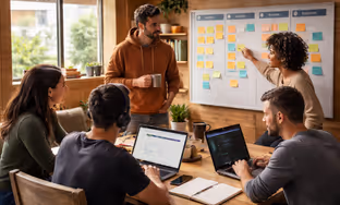Four colleagues collaborate in a bright office, with one pointing at a whiteboard covered in colorful sticky notes while others work on laptops.