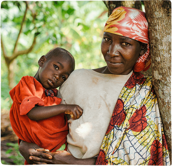A Burundian mother holds her young child while leaning against a tree in front of a lush green background in Burundi
