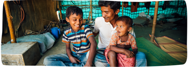 A father smiles at his young son and holds his infant daughter while sitting in their shelter in a Rohingya refugee camp in Bangladesh
