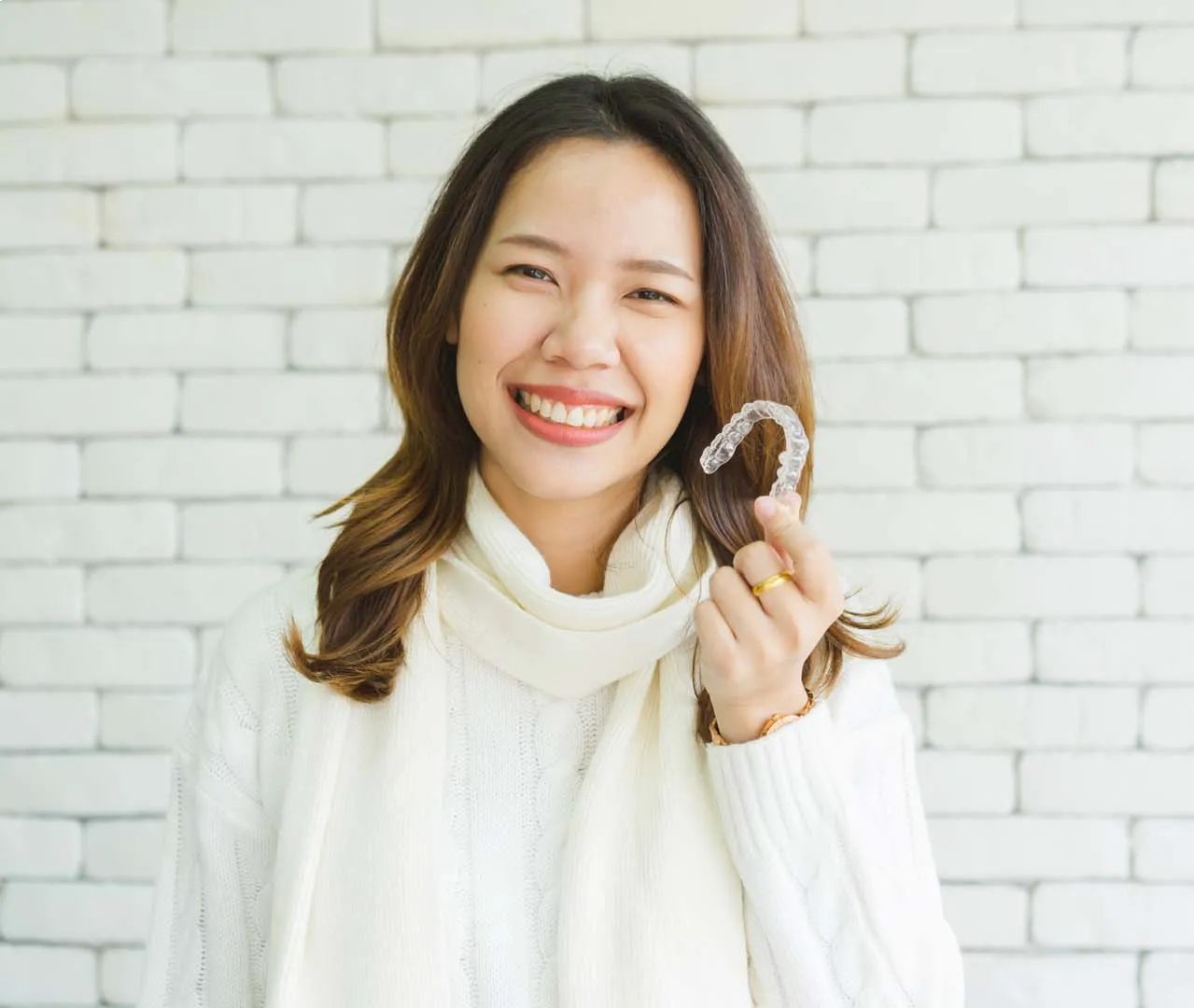 Smiling woman in white sweater holding a clear dental aligner in front of a white brick wall.