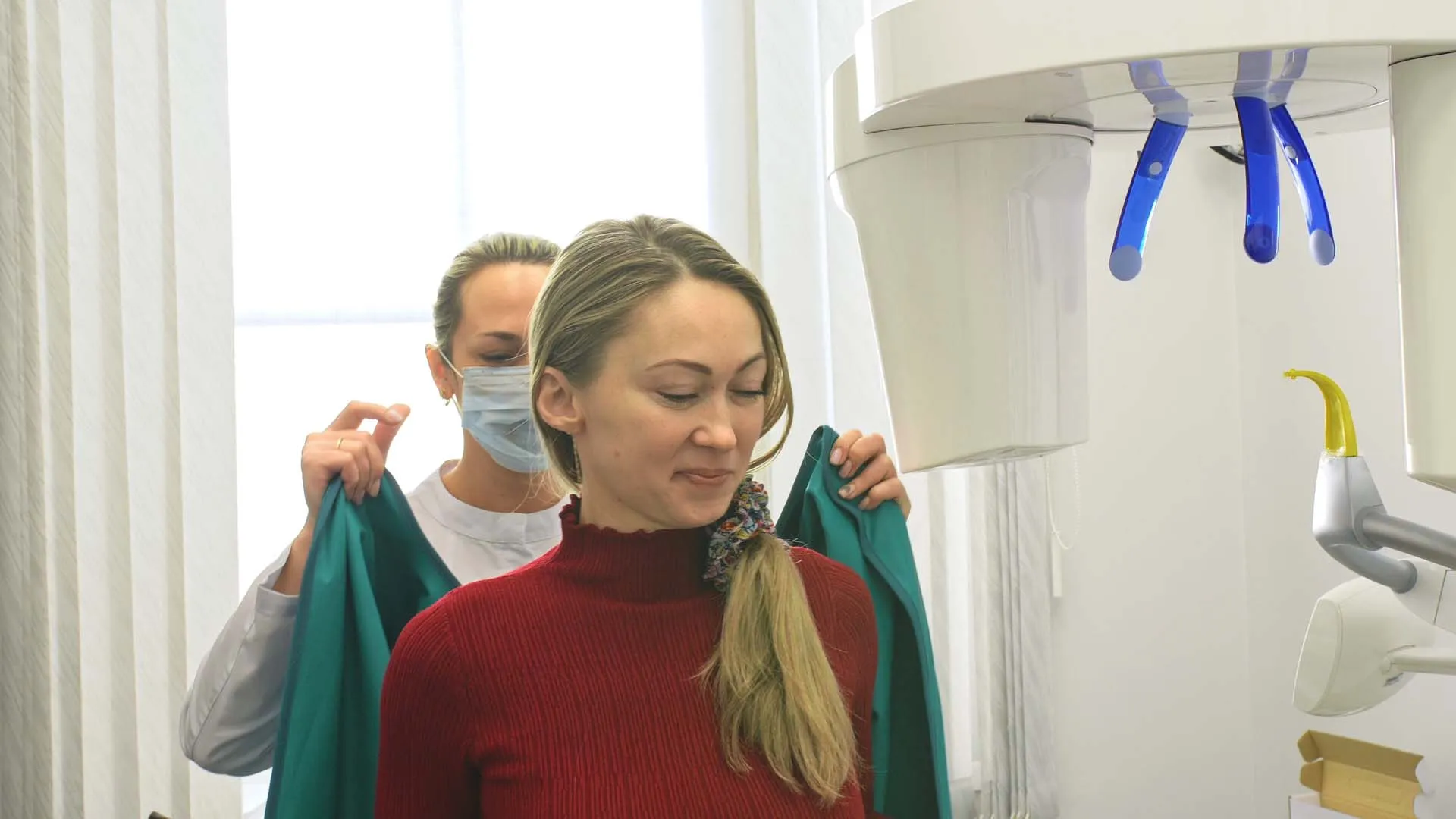 Woman in red sweater being assisted by a masked dental staff with a lead apron near dental X-ray machine.